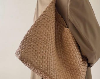 May include: A large, woven, tan leather tote bag with a single strap. The bag is being held by a person wearing a tan jacket.