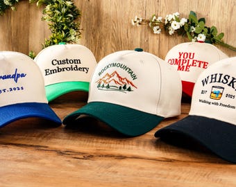 May include: Several baseball caps with embroidered designs. The caps feature various colour schemes, including blue, green, and red peaks. Text on the caps includes "Grandpa", "Custom Embroidery", "Rocky Mountain", "You Complete Me", and "Whisky".