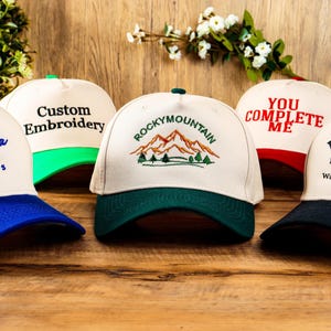 May include: Five different baseball caps with various embroidered designs. The caps are white with different coloured brims. The designs include "Grandpa Est. 2023", "Custom Embroidery", "Rocky Mountain", "You Complete Me", and "Whisky Est. 2023 Walking with Freedom".
