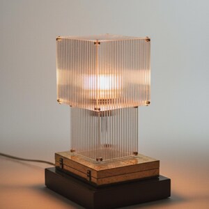 May include: A contemporary table lamp featuring a clear, ribbed, cube-shaped shade. The lamp has a rectangular wooden base and a smaller wooden box in the middle. The lamp is lit, emitting a warm glow. The lamp is approximately 30 cm tall.