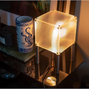 May include: A contemporary cube table lamp emitting a warm glow. The lamp has a clear, ribbed shade and is supported by four silver legs. Other items on the table include a blue and white ceramic vase and a small black container.
