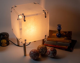 May include: A cube-shaped table lamp with a warm glow, constructed from translucent panels and metal hardware. The lamp sits on wooden legs. Decorative spheres and a small blue glass lamp are nearby.