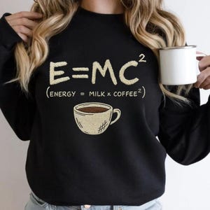 May include: Black sweatshirt featuring the equation E=MC² and the text "(Energy = Milk x Coffee²)". A coffee cup graphic is below the equation. A white mug is held in the person's hand.