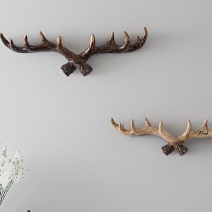 Könnte beinhalten: Zwei geweihförmige Wandhaken. Einer ist dunkelbraun, der andere hellbraun mit dem Namen "Noah" darauf. Die Haken sind an einer hellgrauen Wand angebracht, was auf eine rustikale oder Lodge-Stil-Dekoration hindeutet.