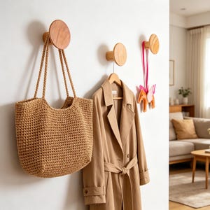 Könnte beinhalten: An einer weißen Wand sind Holzhaken mit natürlichem Finish angebracht. Eine gewebte Tragetasche, ein beigefarbener Trenchcoat an einem Holzkleiderbügel und ein dekoratives Einhorn hängen an den Haken. Der Raum hat eine neutrale Farbpalette.