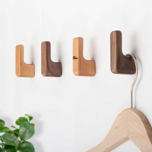 May include: Four wooden wall hooks in varying shades of brown and tan are mounted on a white wall. One hook holds a wooden hanger. The hooks are L-shaped, with a small logo on one of the hooks.
