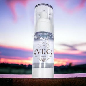 Puede incluir: Una botella cilíndrica transparente con un dispensador de bomba blanco y una tapa transparente. La etiqueta dice "VIVA KOUTOUR" y "VKC COSMETICS" en una fuente decorativa. La botella está llena de un líquido transparente, con un fondo de puesta de sol.