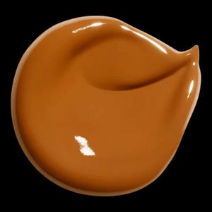 May include: A swatch of liquid foundation in a warm, medium brown shade. The product is smooth and glossy, with a slight sheen, against a black background. The foundation appears to be freshly applied, with a rounded shape and a small peak.