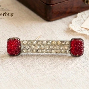Peut inclure: Épingle à nourrice vintage de forme rectangulaire. La broche présente deux pierres rouges texturées et rectangulaires à chaque extrémité. Le centre de l'épingle est une rangée de strass ronds et transparents. Le texte "luluglitterbug EST. 2006" est en haut à gauche.