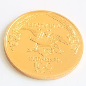Può includere: Una moneta commemorativa color oro con il logo Budweiser, raffigurante un'aquila e la scritta "Budweiser 100 Years" e gli anni 1876-1976. La moneta è di forma circolare e posta su sfondo bianco.