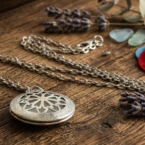 May include: A silver-colored, round pendant necklace with a floral design, resting on a wooden surface. The necklace chain is also silver. Dried lavender and colorful felt circles are in the background.