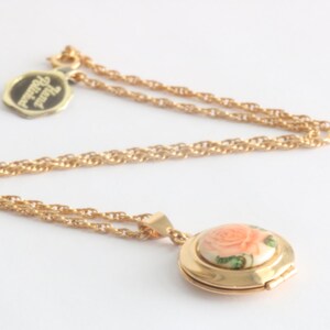 May include: Gold-toned necklace with a pendant featuring a painted rose design. The pendant is round and opens to reveal a small space. A small tag with text hangs near the chain.