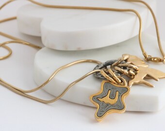 Könnte beinhalten: Eine goldfarbene Halskette mit mehreren Anhängern. Die Anhänger umfassen einen sternförmigen Anhänger mit einem gold-grauen Design, einen Vogel und eine Ansammlung von Gold- und Silberelementen. Die Halskette wird auf einer weißen Oberfläche präsentiert.