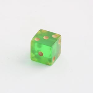 May include: A single, translucent green die with brown dots. The die is a cube shape, with each face displaying a different number of dots. The dots are arranged in the standard pattern for dice, with the numbers one through six.