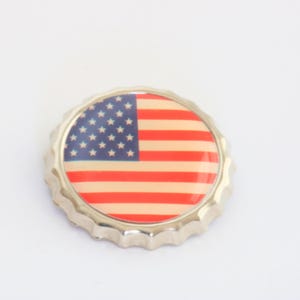 May include: A silver bottle cap with a printed image of the American flag. The flag design features red and white stripes and a blue field with white stars. The cap has a crimped edge.