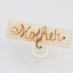 以下が含まれることがあります： 白い長方形のピンに、金色のワイヤーで「母」の文字が書かれています。ピンには、小さな白い丸いチャームがぶら下がっています。