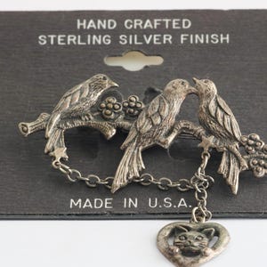Puede incluir: Broche plateada con tres pájaros posados en una rama con bayas. Los pájaros se miran y tienen una cadena con un dije en forma de corazón con la cara de un gato que cuelga de la rama. La tarjeta dice "Hand Crafted Sterling Silver Finish" y "Made in U.S.A."