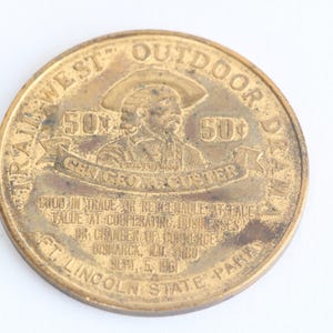 May include: A round, antique-looking bronze coin with the text "TRAIL WEST OUTDOOR DAYS" and "GEORGE A. CUSTER". The coin features a portrait of a man in a hat and the text "GOOD IN TRADE OR REDEEMABLE AT FACE VALUE".