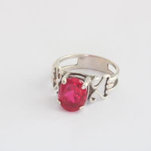 May include: A silver ring featuring a large, oval-cut, bright red gemstone. The ring has an openwork design on the sides, with decorative cutouts. The gemstone is the focal point, set in a classic prong setting.