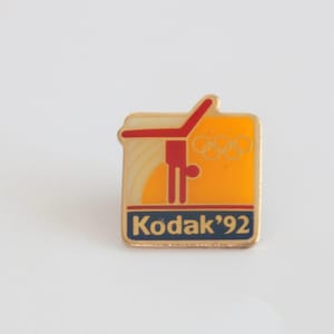 May include: A vintage Kodak '92 Olympic pin. The pin features a gold border, a yellow background with a red gymnast, and the Olympic rings. The text "Kodak '92" is in a blue rectangle.