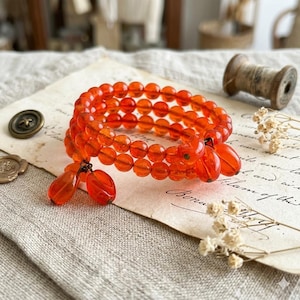 May include: An orange beaded bracelet with three strands of round beads. The bracelet has three teardrop-shaped charms. The bracelet is resting on a vintage paper with a button and a spool of thread in the background.