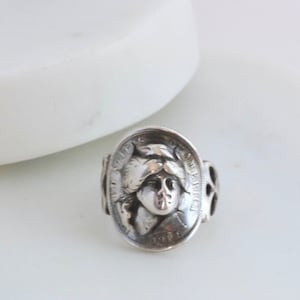 Może przedstawiać: Srebrny pierścionek z vintage'ową monetą z 1917 roku, Walking Liberty Half Dollar. Moneta jest osadzona w srebrnej obręczy z ażurowym wzorem.