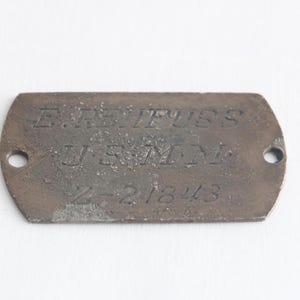 May include: A weathered, rectangular metal dog tag with rounded edges. The tag has two holes and features the engraved text "E. REIFUSS", "U.S.M.M.", and "Z-21843". The metal has a dark, aged appearance.
