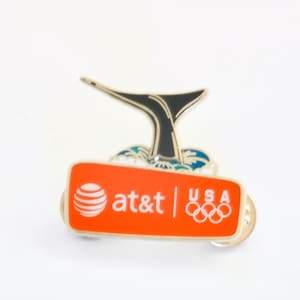 May include: A pin featuring a black whale tail rising from blue and white waves. Below, an orange rectangle displays the AT&T logo and the Olympic rings with "USA" text. The pin has a silver border.