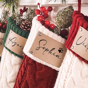 May include: Four cable-knit Christmas stockings in red, green, and cream colors. Each stocking features a tan leather-like rectangular patch attached with four silver rivets. The names Janice, Agnes, Sophia, and Victoria are printed on the patches.