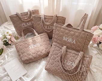 Puede incluir: Una colección de cinco bolsos tote acolchados con un patrón de cuadros vichy en marrón y blanco. Cada bolso tiene dos asas y está bordado con un nombre único en hilo blanco, concretamente Clara, Ollie, Nora, Finn y Liza.