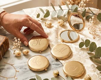 May include: Four round gold-toned metal compact mirrors, each engraved with a different cursive letter: K, M, C, and S. One mirror is shown open, revealing a reflective surface, while the others are closed, displaying their engraved monograms.