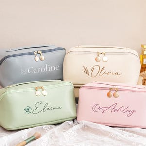 Peut inclure: Quatre trousses de maquillage personnalisées aux couleurs pastel : bleu clair, crème, vert et rose. Chaque trousse est dotée d'une fermeture éclair dorée et d'un nom brodé dans une couleur complémentaire. Les noms sont Caroline, Olivia, Elaine et Ashley.