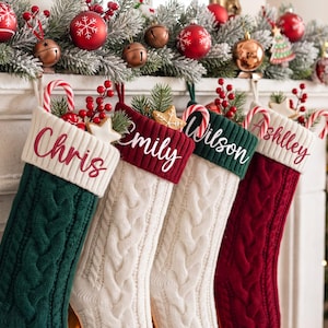 Könnte beinhalten: Vier Weihnachtsstrümpfe aus Zopfstrick in Dunkelgrün, Creme und Dunkelrot, jeweils mit weißem Rippbündchen, personalisiert mit den Namen Chris, Emily, Wilson und Ashlley, gefüllt mit Zuckerstangen, roten Beeren und sternförmigen Keksen.