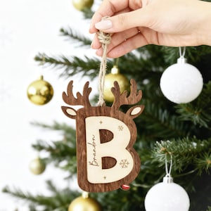 以下が含まれることがあります： トナカイの頭の形をした木製のクリスマスオーナメント。文字「B」と「Brandon」の名前が刻まれています。オーナメントは茶色で、明るい木材のアクセントと紐のループが付いています。背景には、オーナメントが付いたクリスマスツリーが写っています。