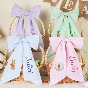 May include: Four Easter baskets with large striped bows in pastel colours: purple, green, pink, and blue. Each bow has a name embroidered on it. The baskets have embroidered bunny designs and are filled with Easter eggs and carrots.