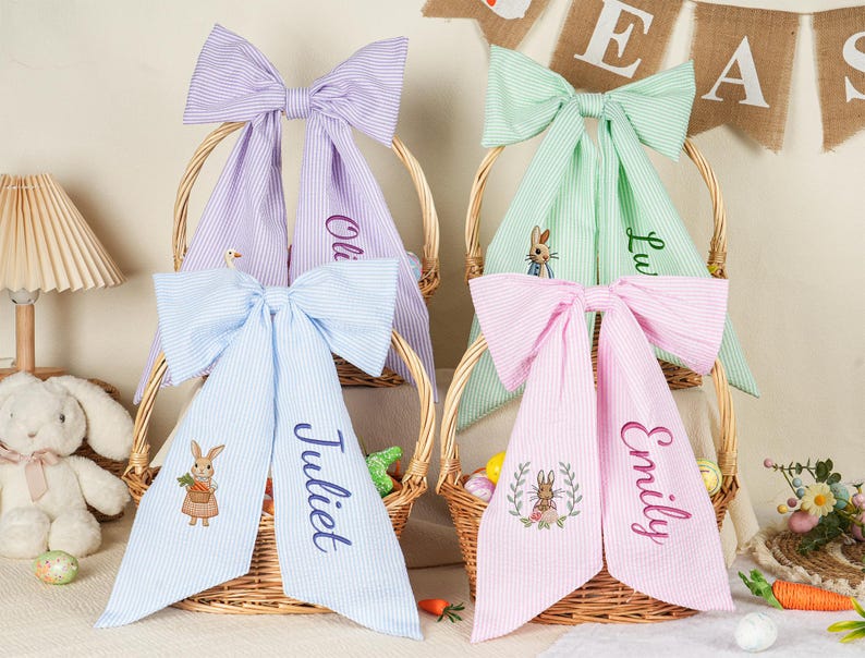 Pode incluir: Quatro cestas de P&aacute;scoa com grandes la&ccedil;os listrados em cores pastel: roxo, verde, rosa e azul. Cada la&ccedil;o tem um nome bordado. As cestas t&ecirc;m desenhos de coelhinhos bordados e s&atilde;o preenchidas com ovos e cenouras de P&aacute;scoa.