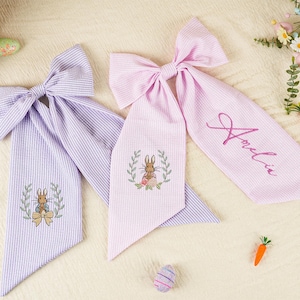 Pode incluir: Duas fitas de cabelo em tecido seersucker, uma lavanda e outra rosa, com desenhos de coelhos bordados e o nome "Amelia" em uma. As fitas s&atilde;o decoradas com elementos tem&aacute;ticos da P&aacute;scoa, como cenouras e ovos.