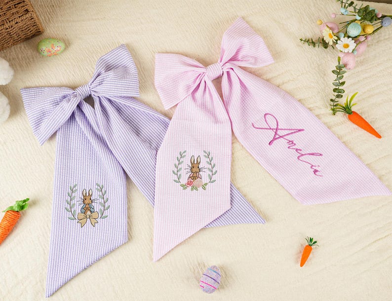 Pode incluir: Duas fitas de cabelo em tecido seersucker, uma lavanda e outra rosa, com desenhos de coelhos bordados e o nome "Amelia" em uma. As fitas s&atilde;o decoradas com elementos tem&aacute;ticos da P&aacute;scoa, como cenouras e ovos.