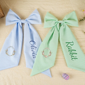 Pode incluir: Duas fitas de cabelo com tema de P&aacute;scoa. Uma &eacute; azul clara com o nome "Olivia" bordado, e a outra &eacute; verde com "Rabbit". Ambas t&ecirc;m um desenho de coelho e grinalda. Tamb&eacute;m est&atilde;o presentes ovos e cenouras decorativas de P&aacute;scoa.