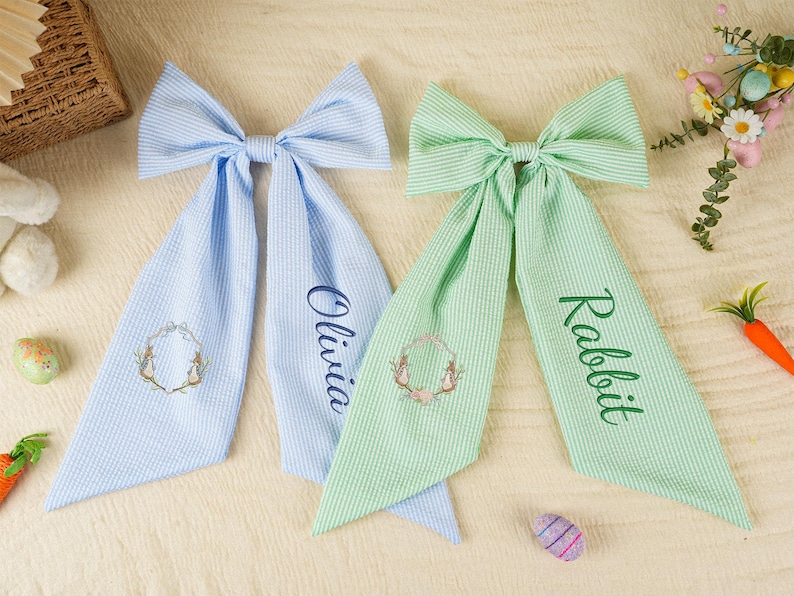 Pode incluir: Duas fitas de cabelo com tema de P&aacute;scoa. Uma &eacute; azul clara com o nome "Olivia" bordado, e a outra &eacute; verde com "Rabbit". Ambas t&ecirc;m um desenho de coelho e grinalda. Tamb&eacute;m est&atilde;o presentes ovos e cenouras decorativas de P&aacute;scoa.
