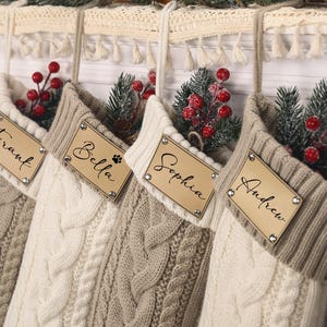 Może przedstawiać: Cztery świąteczne skarpety w warkoczowy wzór w odcieniach kremu i beżu wiszą na kominku. Każda skarpeta ma drewnianą metkę z imieniem i jest ozdobiona sztucznymi jagodami i zielenią.