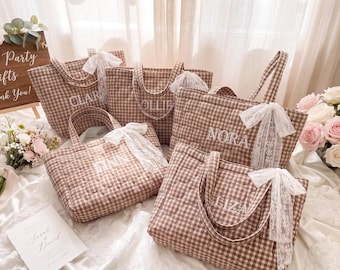 May include: Four quilted tote bags in a brown and white gingham pattern, each featuring a white embroidered name and a white lace bow attached to the handle. The names embroidered on the bags are Clara, Ollie, Nora, and Liza.