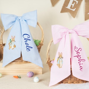 Pode incluir: Dois la&ccedil;os para cestas de P&aacute;scoa, um azul claro e outro rosa, com listras seersucker. Cada la&ccedil;o tem um nome personalizado bordado nas caudas. O la&ccedil;o azul tem "Chloe" bordado, e o la&ccedil;o rosa tem "Sophia". Ambos os la&ccedil;os apresentam um coelho bordado.