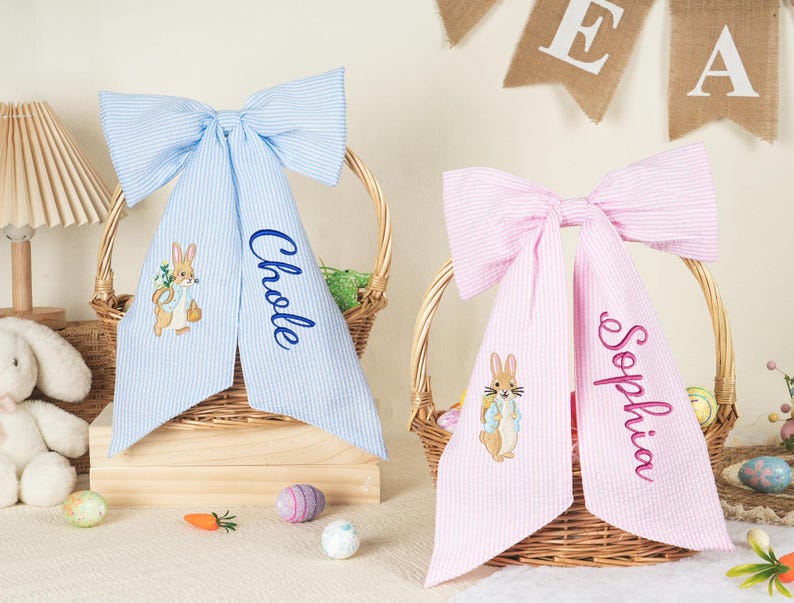 Pode incluir: Dois la&ccedil;os para cestas de P&aacute;scoa, um azul claro e outro rosa, com listras seersucker. Cada la&ccedil;o tem um nome personalizado bordado nas caudas. O la&ccedil;o azul tem "Chloe" bordado, e o la&ccedil;o rosa tem "Sophia". Ambos os la&ccedil;os apresentam um coelho bordado.