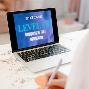 Puede incluir: Una pantalla de computadora portátil que muestra un tutorial sobre hielo seco para la presentación de mesas de bebidas y postres. La pantalla dice "Dry Ice Tutorial" y "Level Up" en texto azul. El texto "Drink/Dessert Table Presentation" está en texto blanco. Una persona está escribiendo en un papel rojo en forma de corazón.