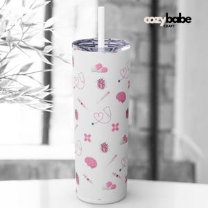 May include: White stainless steel tumbler with a pink and white pattern of medical symbols, including hearts, pills, stethoscopes, brains, and syringes. The tumbler has a lid and a straw.