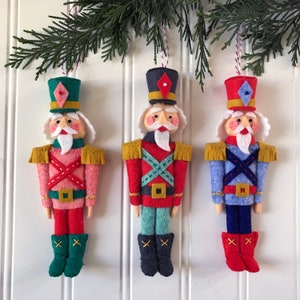 Peut inclure: Trois ornements de Noël faits à la main en feutre en forme de soldats casse-noisettes. Chaque soldat a une combinaison de couleurs différente : rose, rouge et bleu. Ils ont des barbes blanches, des épaulettes dorées et des chapeaux décoratifs. Les ornements sont suspendus à une branche de pin verte.