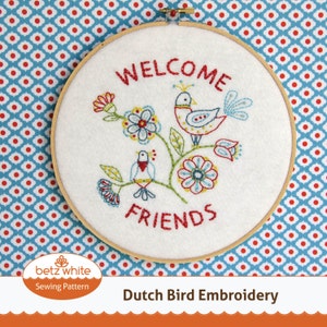 May include: A white fabric embroidery hoop with the words "WELCOME FRIENDS" stitched in red thread. The design features colorful embroidered birds and flowers. The wooden hoop is set against a blue and white patterned background.