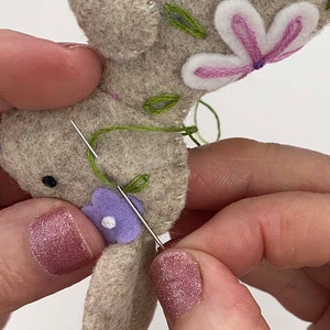 May include: Close-up of a felt bunny being hand-stitched with green and purple thread. The bunny is gray with white and purple flower accents. A needle is inserted through the felt, showing the embroidery process.