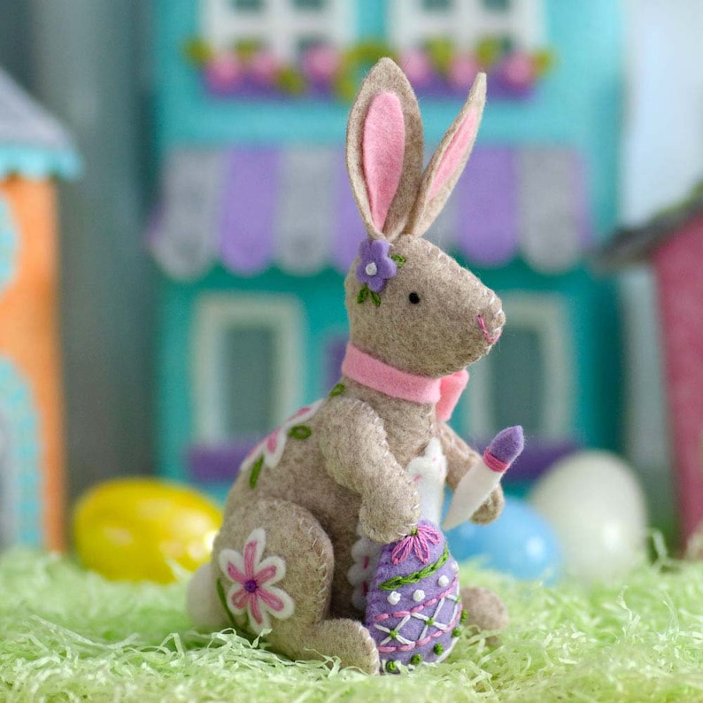 May include: A tan felt Easter bunny figurine with pink-lined ears, a pink scarf, and a purple flower. The bunny holds a decorated purple egg and a paintbrush. White and pink flowers decorate the bunny's body. The bunny is sitting on green paper grass.
